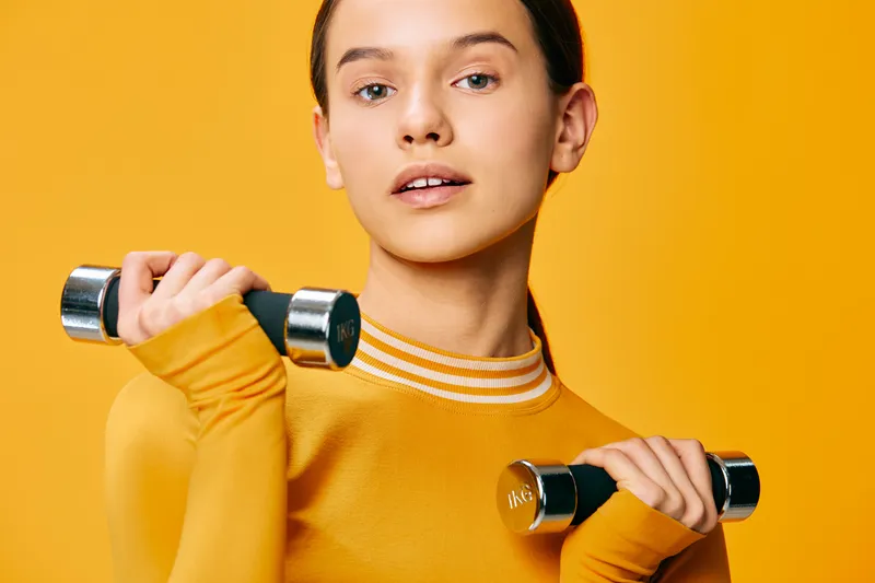 Woman fitness, sport, lifting dumbbells in a bright yellow top against an orange backdrop, performing strength training, focused exercise routine and healthy active lifestyle.