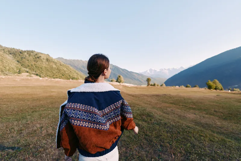 Woman in a cozy sweater walking across a mountain meadow, nature landscape with scenic outdoors scenery. Back view portrait captures calm hiking journey and tranquil horizon.