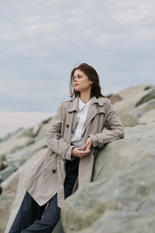 Young woman in a long coat leans on large rocks outdoors under cloudy sky. Concept of autumn fashion, natural beauty, and thoughtful mood in serene coastal environment.