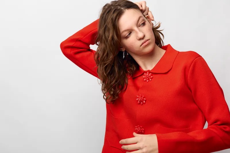 Young woman with curly brown hair wearing a red sweater looks away thoughtfully, hand touching head. Studio portrait on white background with casual fashion and natural expression.
