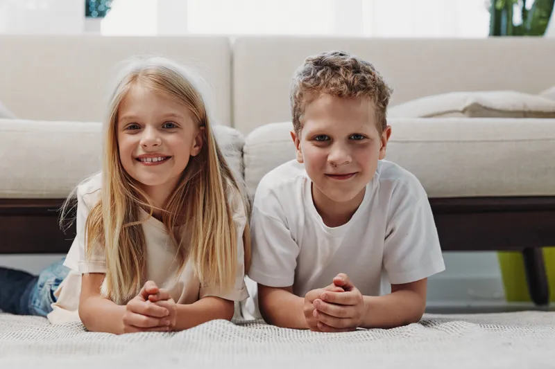 Happy girl and boy lying on carpet in living room, smiling at camera. Casual children in white shirts, cozy home environment with sofa and natural light. Concept of childhood, friendship, family.