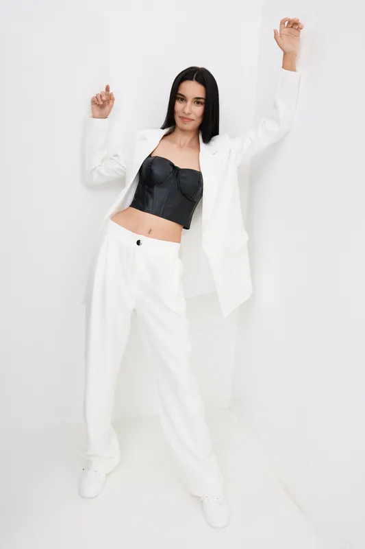 stylish woman wearing white pantsuit and black corset top posing confidently against a white wall, showcasing modern fashion and minimalistic background in bright lighting