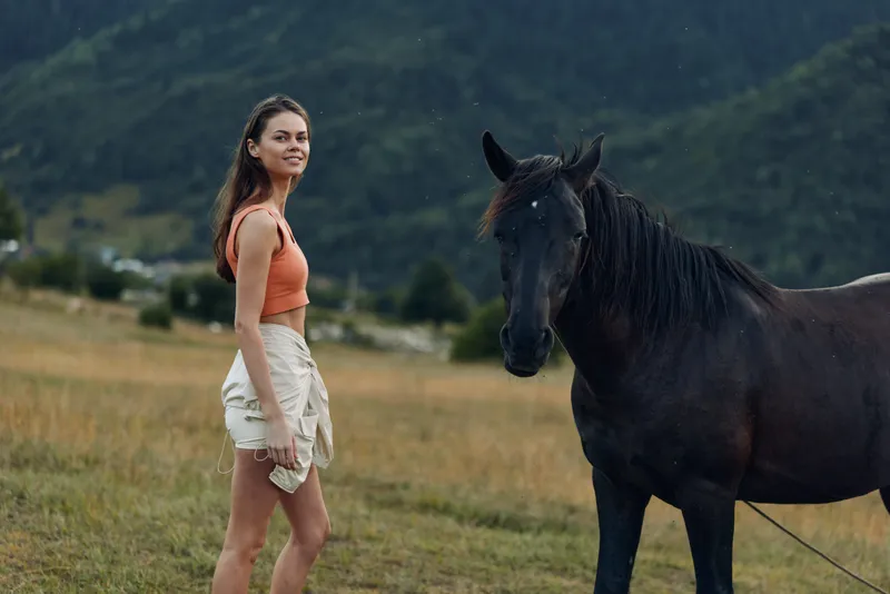 A cheerful woman stands in a grassy field with a dark horse beside her. She smiles warmly, wearing a light top and shorts, enjoying a tranquil outdoor moment.