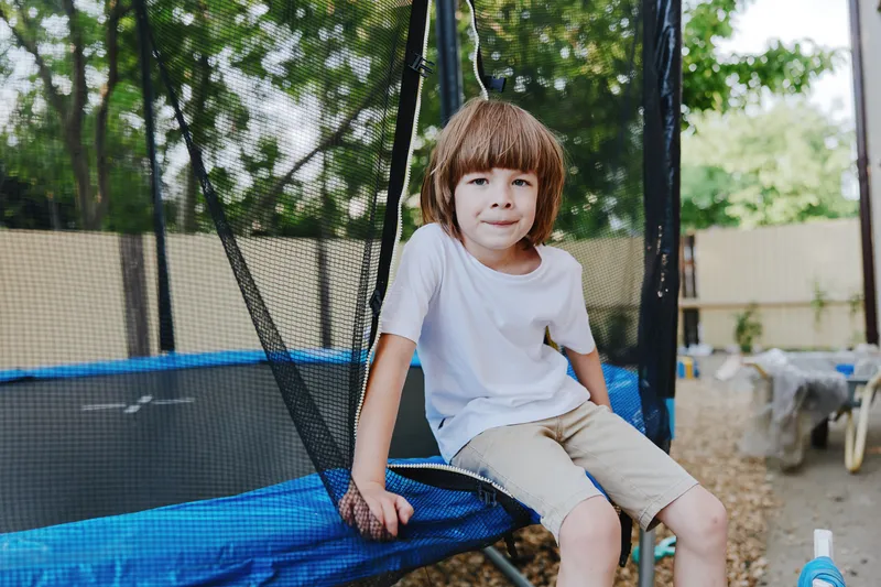 Playful boy enjoying a sunny day, sitting on a trampoline, surrounded by greenery and outdoor fun, reflecting joy and carefree childhood moments.