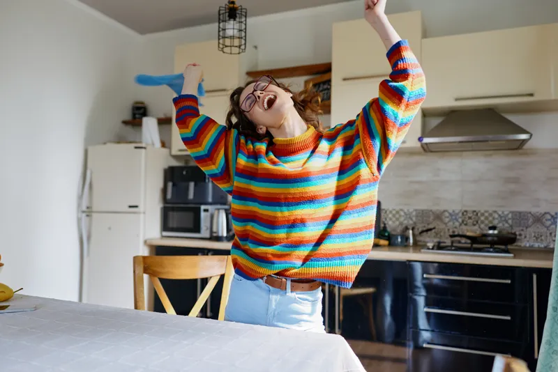 Cheerful woman indoors, dancing in the kitchen, wearing a colorful striped sweater, casual lifestyle moment, joyful and carefree, bright morning light, warm atmosphere, cozy home setting