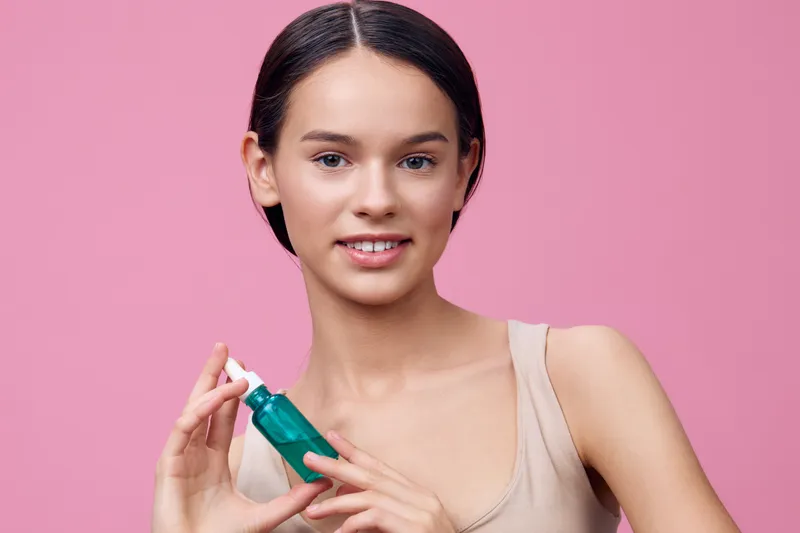 Young woman with clear skin holding a green skincare bottle, smiling against a pink background, representing beauty and self care