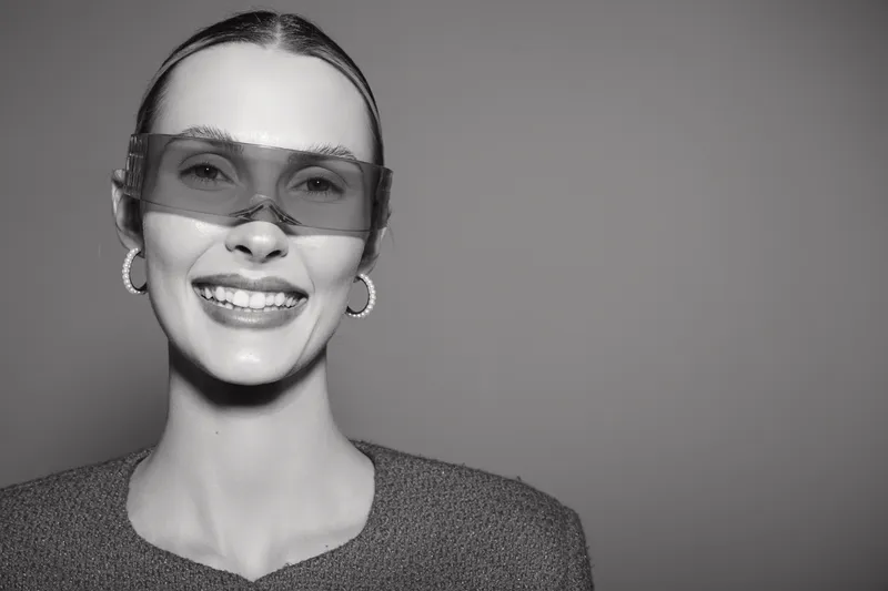 Smiling white woman with pale skin wearing transparent sunglasses and hoop earrings. Studio portrait in black and white with smooth hairstyle and textured top. Concept of fashion and happiness.