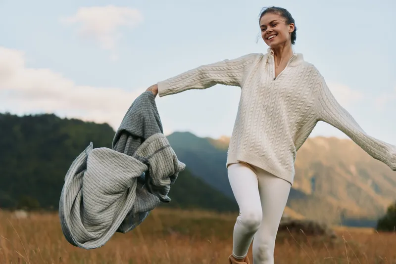 Cheerful lifestyle moment of a woman outdoors, laughing with eyes closed, standing in a meadow with a blanket, distant mountains behind, bright sunlight and a soft breeze
