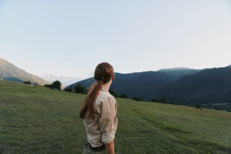A young woman in a beige shirt stands in a green hillside meadow, gazing toward distant mountains, conveying solitude and contemplation.