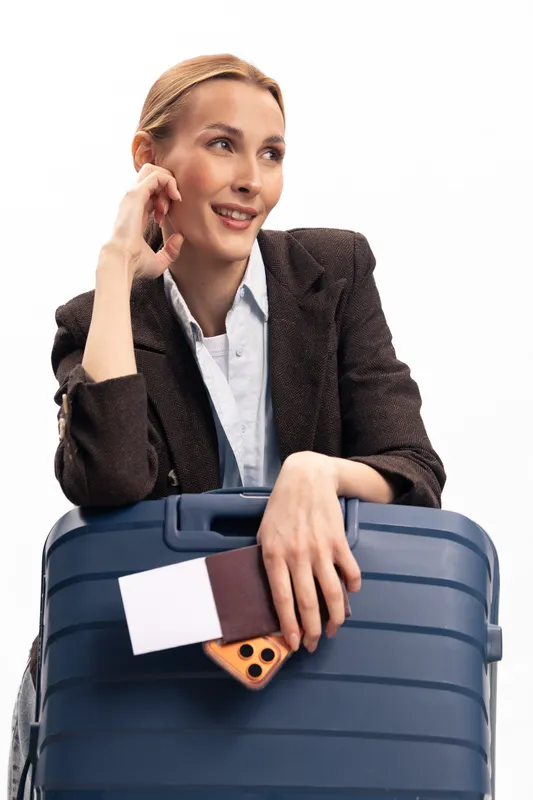 Smiling woman in brown jacket leans on blue suitcase holding passport and smartphone. Travel concept, airport preparation, vacation planning, and holiday anticipation on white background.