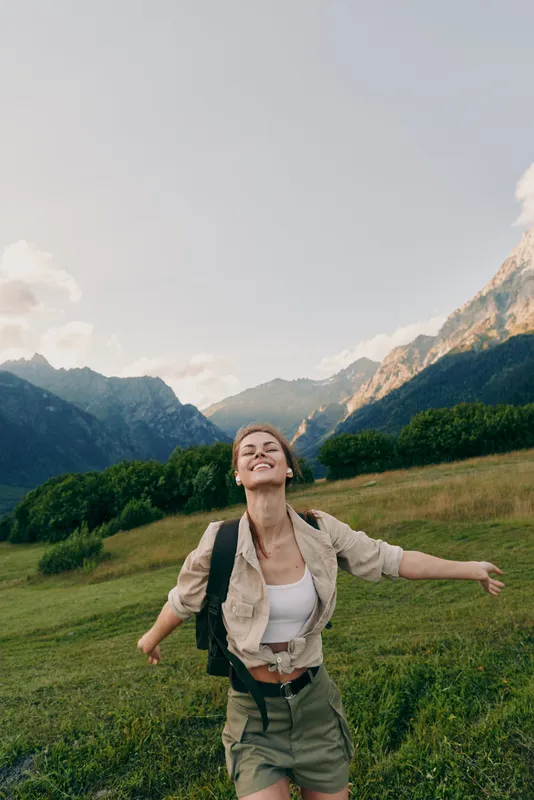 Joyful woman hiking in a green meadow with mountains in the background, wearing a light jacket and backpack. Outdoor adventure and wellness. Embracing nature Outdoors.