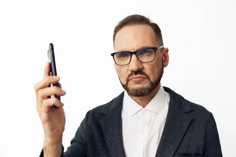 A professional man in a blazer with glasses holds a smartphone, posed against an isolated background. He conveys focus, seriousness, and quiet determination in business.