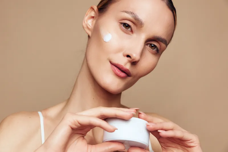 Young woman with clear skin applies moisturizer cream on her face while holding a white jar against a plain background in a skincare routine moment.