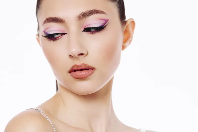 Close-up of young woman with creative bright pink and purple eye makeup standing against white background. Concept for beauty, fashion, makeup trends, and artistic expression.