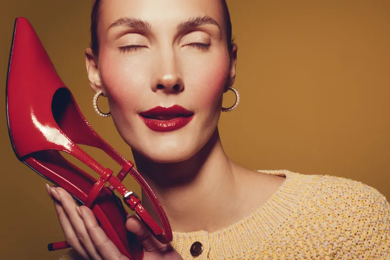 Close-up of a woman with closed eyes holding a red high heel shoe near her face. Warm lighting highlights her smooth skin and red lipstick on a brown background. Concept of fashion and beauty.
