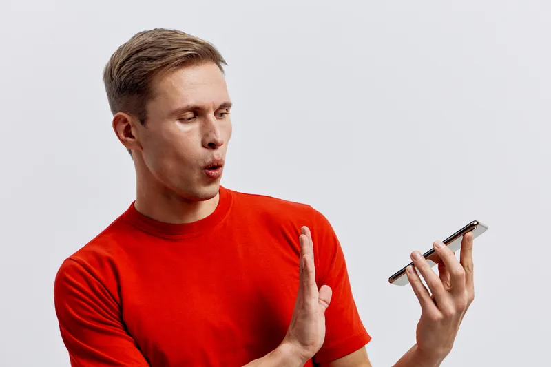 Confused man in casual red T-shirt looking at smartphone with surprised expression isolated on plain white background, studio portrait, lifestyle concept, facial expression