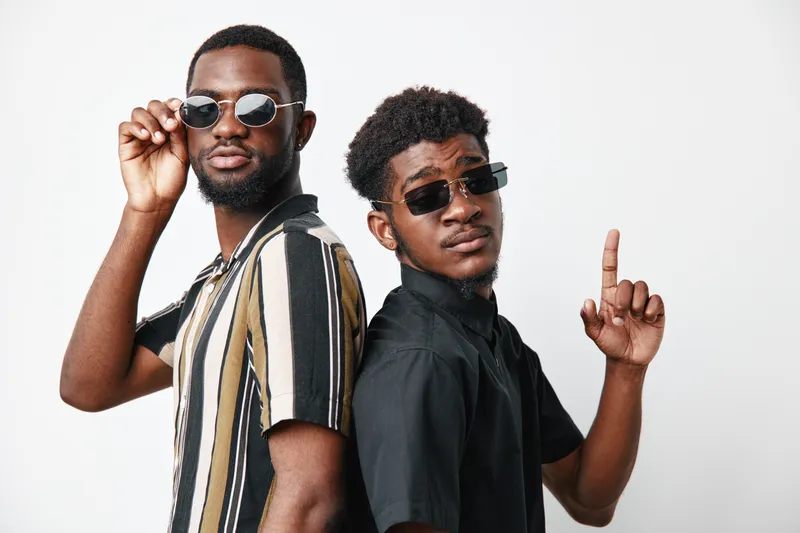 Two young black men with dark skin wearing sunglasses posing confidently against white background. Casual style, striped shirt and black shirt, cool attitude and modern fashion.