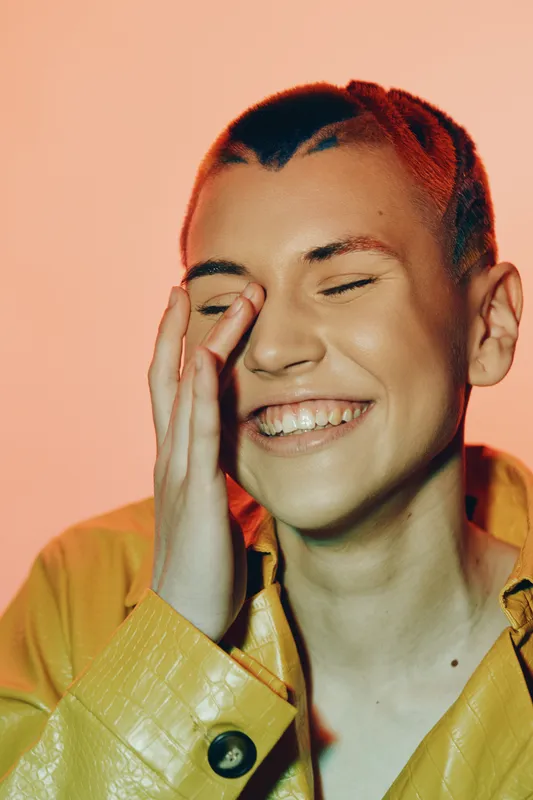 smiling woman with short red hair wearing a yellow jacket laughing joyfully with closed eyes on a warm peach background, capturing happiness and carefree emotion