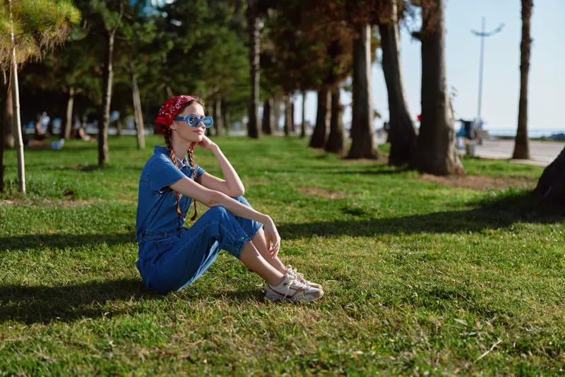 lifestyle woman in modern boho-western denim street style sitting on grass with sunglasses and bandana in warm natural light, film color aesthetic