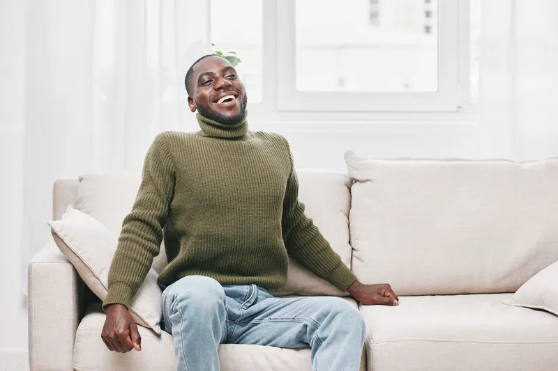 Joyful Black man in a green turtleneck sweater sitting on a light sofa, smiling warmly, conveying happiness and comfort in a bright, airy living room