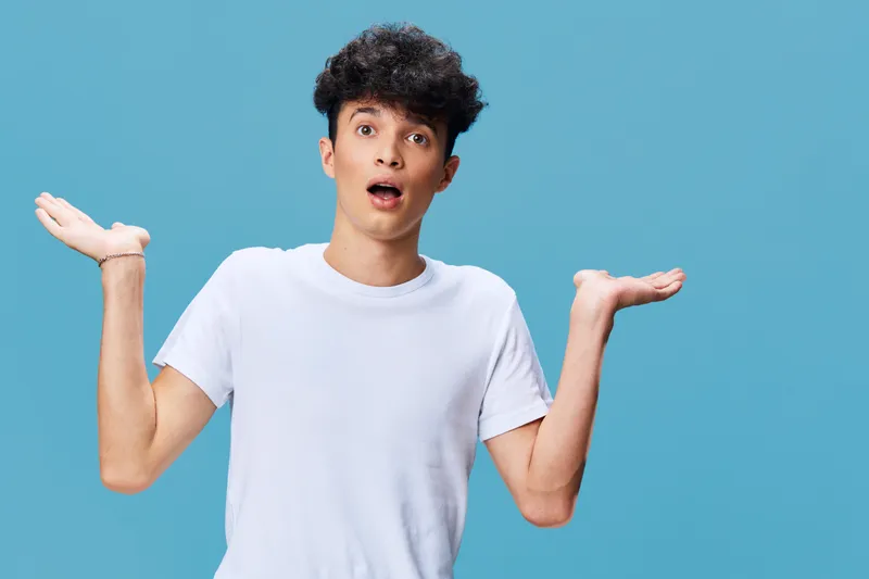 surprised young man with curly hair wearing white t-shirt raising hands against blue background, expressing confusion or disbelief in casual style