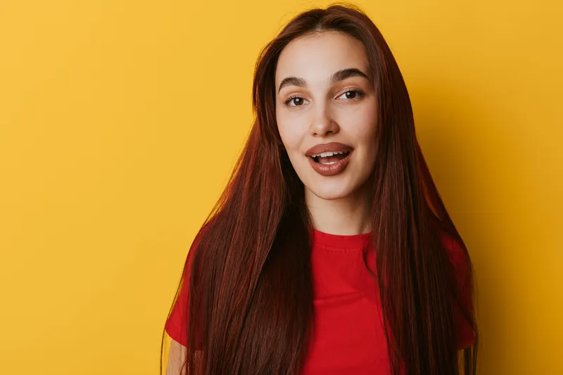 Young woman with long straight hair and red shirt smiling with open mouth standing against bright yellow background in studio. Portrait of happy female with natural makeup and casual style.