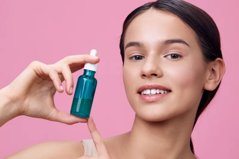 Young woman holding a turquoise serum bottle, smiling confidently against a pink background, showcasing healthy skin and beauty products