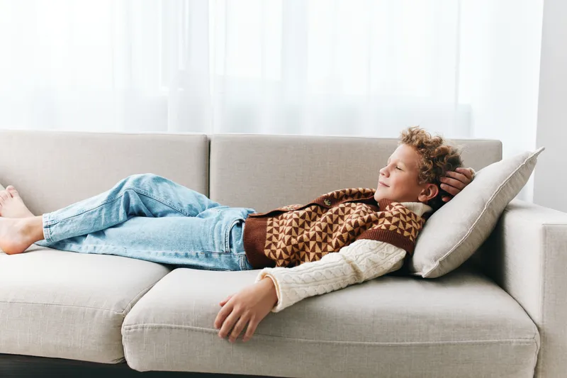 Authentic lifestyle scene showing a student at home, relaxing on a couch, wearing casual knitwear and denim, with playfulness and school life as core themes.
