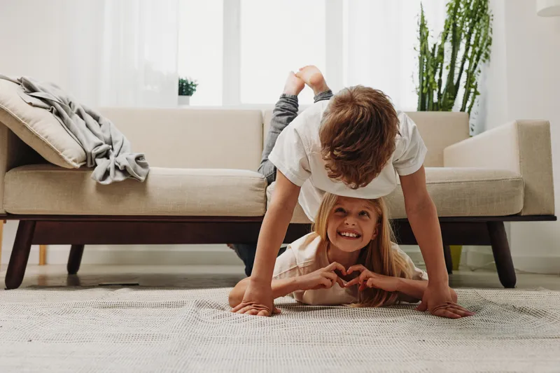 Playful siblings having fun in a bright living room. Boy balances on hands above smiling girl lying on carpet. Concept of family bonding, joy, childhood, and indoor playtime.