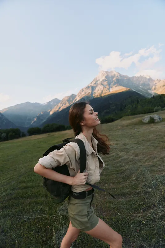 Young woman with a backpack hikes through a grassy meadow toward distant mountains with a clear blue sky and warm sunlight, ready for an adventurous day.