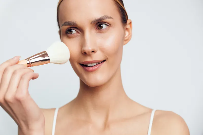 Young woman with clear skin applying powder makeup with a brush on white background. Concept of beauty, skincare, and natural makeup routine with soft lighting and fresh look.