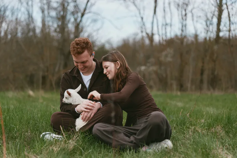 couple goat kid outdoors nature grass affection happiness in a candid lifestyle moment with a man holding a goat and a woman smiling on a grassy field, celebrating companionship and animal care