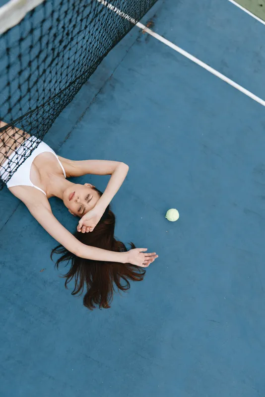 Young woman lying on a tennis court, showcasing a relaxed posture with a tennis ball nearby, emphasizing sports lifestyle and wellness