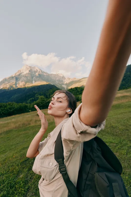 Young woman outdoors raises her arm for a selfie, smiling with eyes closed as the sun lights a grassy hillside with distant mountains, wearing a beige shirt and backpack.