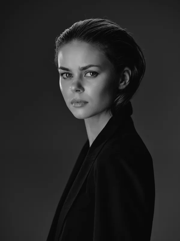 Black and white portrait of a serious woman with slicked-back hair wearing a dark jacket in studio lighting.