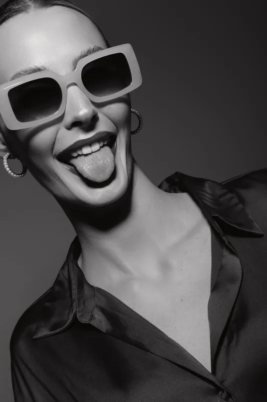 Happy white woman with light skin wearing large sunglasses and silk blouse sticks out tongue in playful studio portrait in black and white.