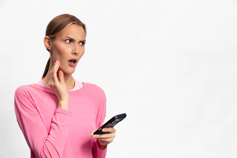 Surprised woman in pink sweater holds a smartphone and looks shocked with open mouth. Isolated on white background. Concept of unexpected news, communication, and emotional reaction.