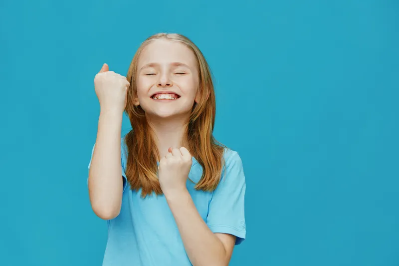 Happy girl with long hair celebrating success in a blue t shirt against a bright blue background, expressing joy with clenched fists and a big smile