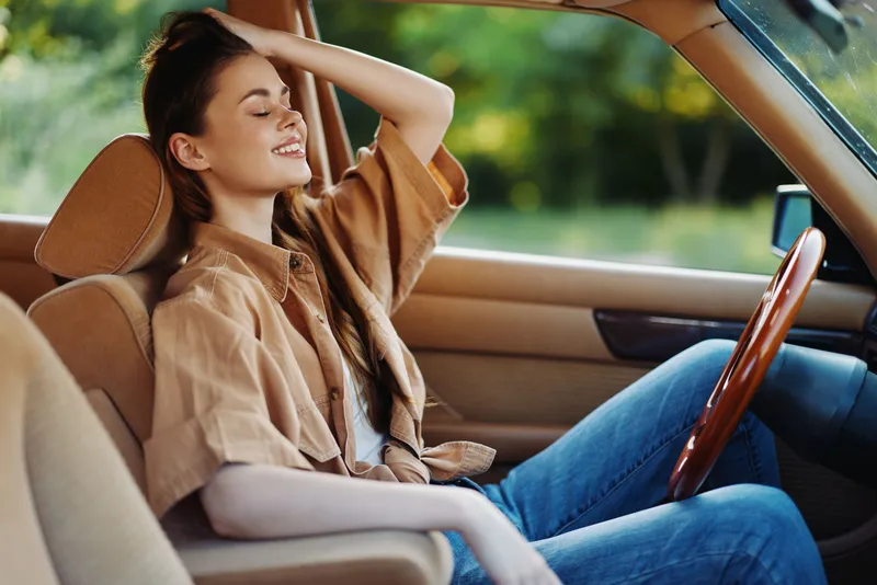 A joyful young woman with long brown hair sitting in a vintage car, wearing a casual beige shirt and jeans, enjoying the warm sunlight in a lush green park.