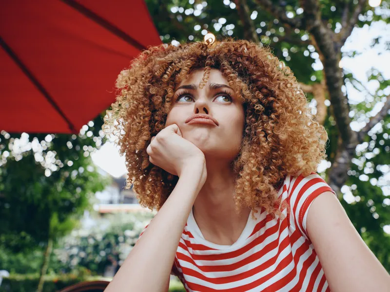Young woman with curly hair rests her chin on her hand beneath a red umbrella, gazing upward with a dreamy, introspective expression in a sunny outdoor garden scene.