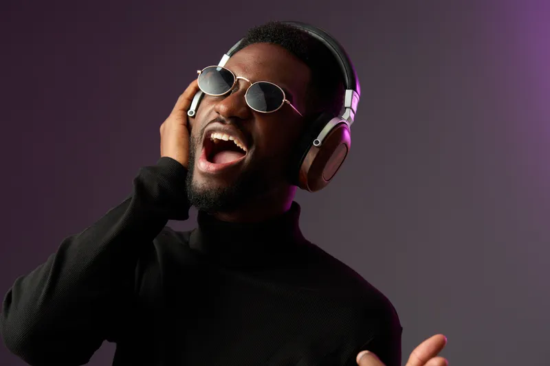Young Black man singing joyfully with headphones, wearing stylish sunglasses against a vibrant gradient background, embodying the joy of music and self expression