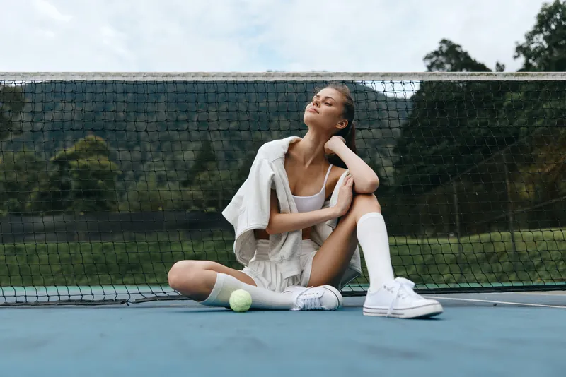 Young female athlete in tennis attire exudes confidence and relaxation while sitting on the court, surrounded by nature, showcasing healthy lifestyle and sportswear