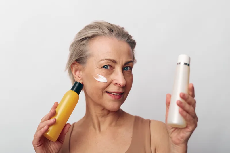 Mature woman with short hair applies cream on face holding two cosmetic bottles on white background. Skincare routine, anti aging care, beauty concept for healthy skin and self care.