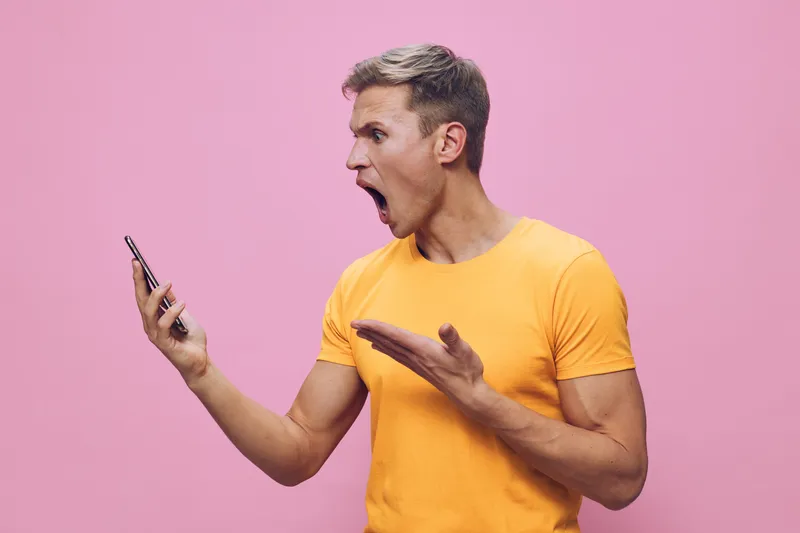 Dynamic young man with short hair wearing bright yellow t-shirt expressing surprise while looking at smartphone in hand, gesturing with other hand, against pink background. People lifestyle concept, e
