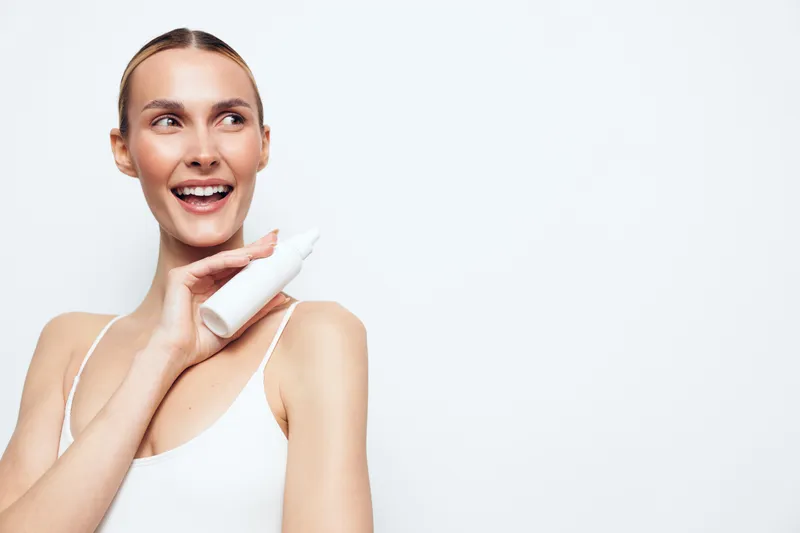 A smiling young woman in a white tank top holds a white cosmetic bottle near her shoulder, looking away. The bright studio background emphasizes skincare and beauty concept.