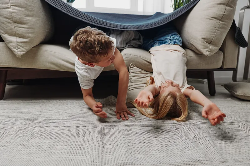 Two playful children crawl under a sofa in a bright living room, engaging in fun activities that showcase childhood joy and sibling bonding in a cozy home environment.