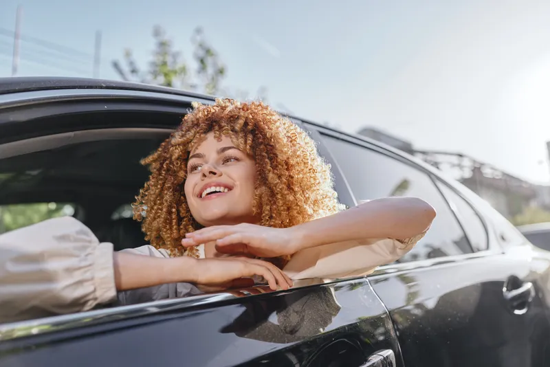 happy woman with curly hair leaning from car window enjoying sunny day outdoors in casual light blouse smiling joyfully