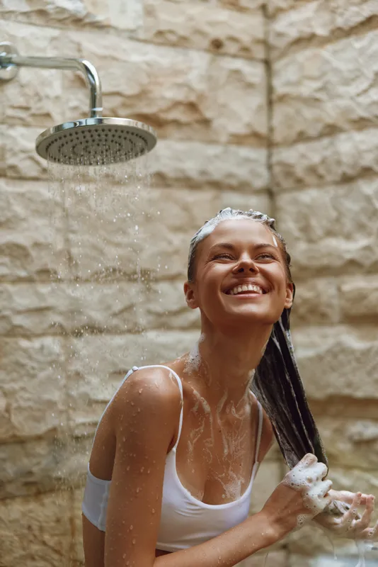 smiling woman washing hair under outdoor shower with stone wall background, fresh water droplets, natural light, healthy skin care, summer wellness moment
