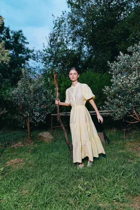 Woman in a pale yellow dress stands in a grassy garden with a wooden staff under a clear blue sky, creating a serene outdoor portrait.