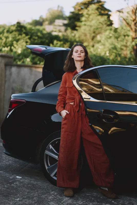 A confident woman in a red coat leans beside a sleek car in an outdoor setting, showcasing contemporary street style and poised lifestyle inspiration